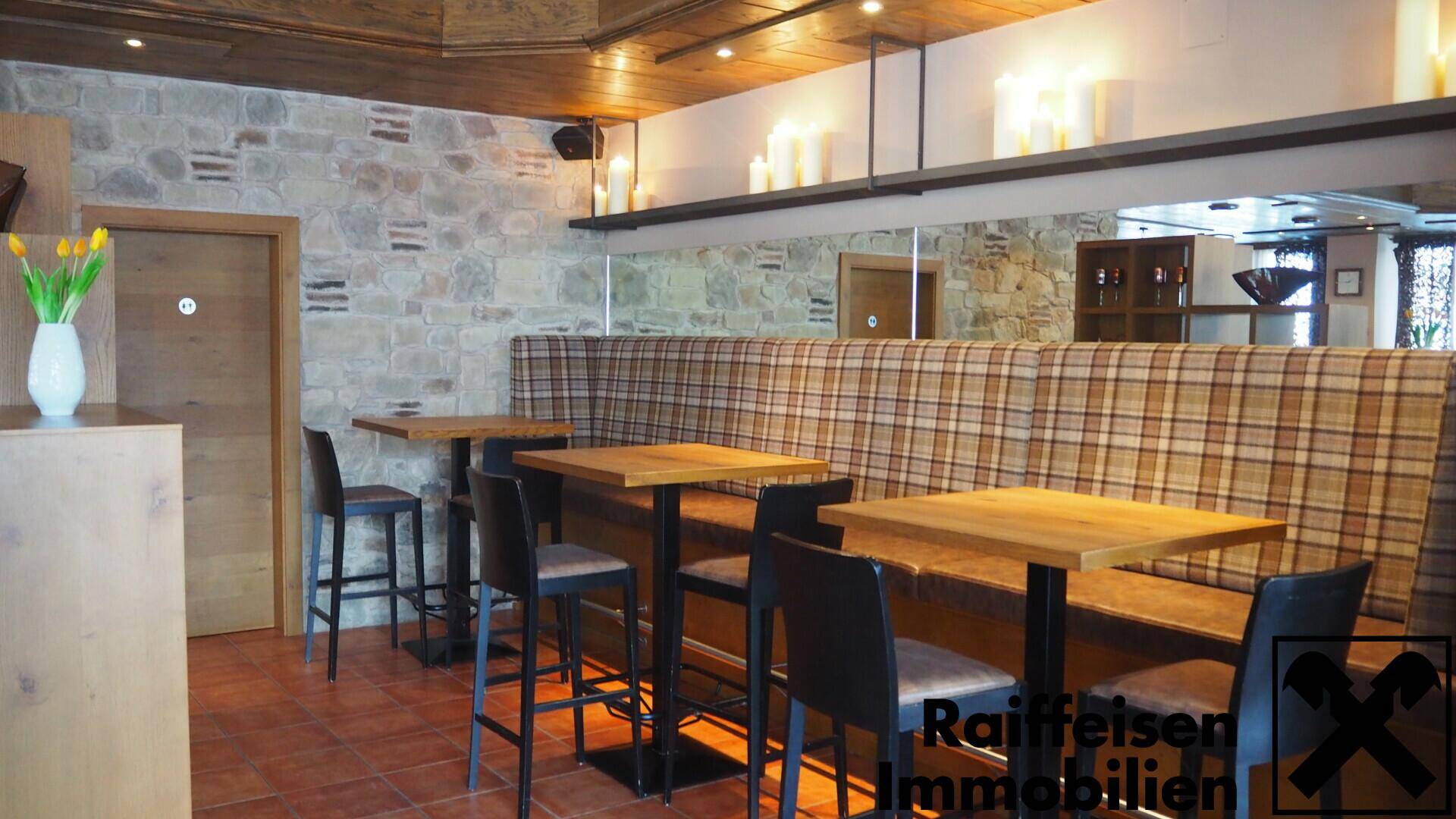 Gemütlicher Restaurantbereich mit kleinen Holztischen, hohen Stühlen und karierten Sitzbänken vor einer Steinwand.