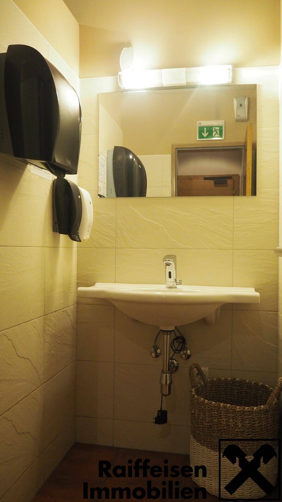 Waschbereich der Toilette mit Waschbecken, Spiegel und Handtrockner, ausgestattet mit hellen Fliesen.
