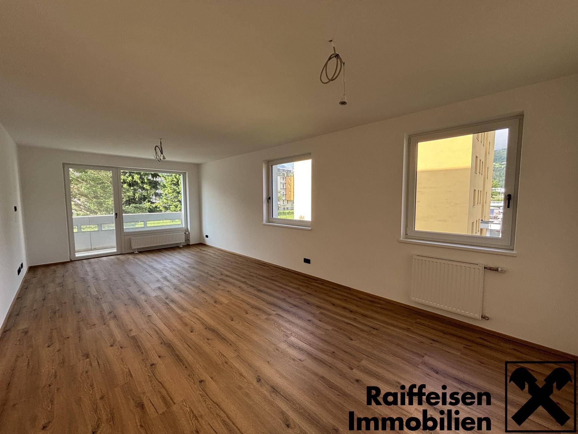 Geräumiges Wohnzimmer mit Laminatboden, großen Fenstern und Zugang zu einem Balkon.