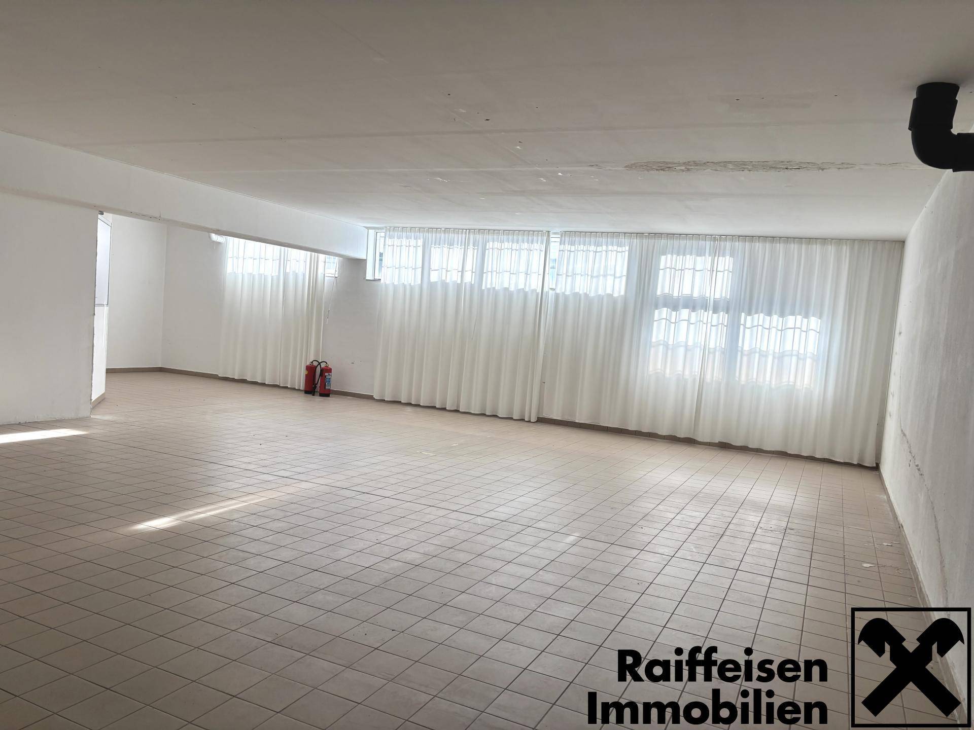 Geräumiger, heller Gewerberaum mit vielen Fenstern, weißen Vorhängen und beigem Fliesenboden.