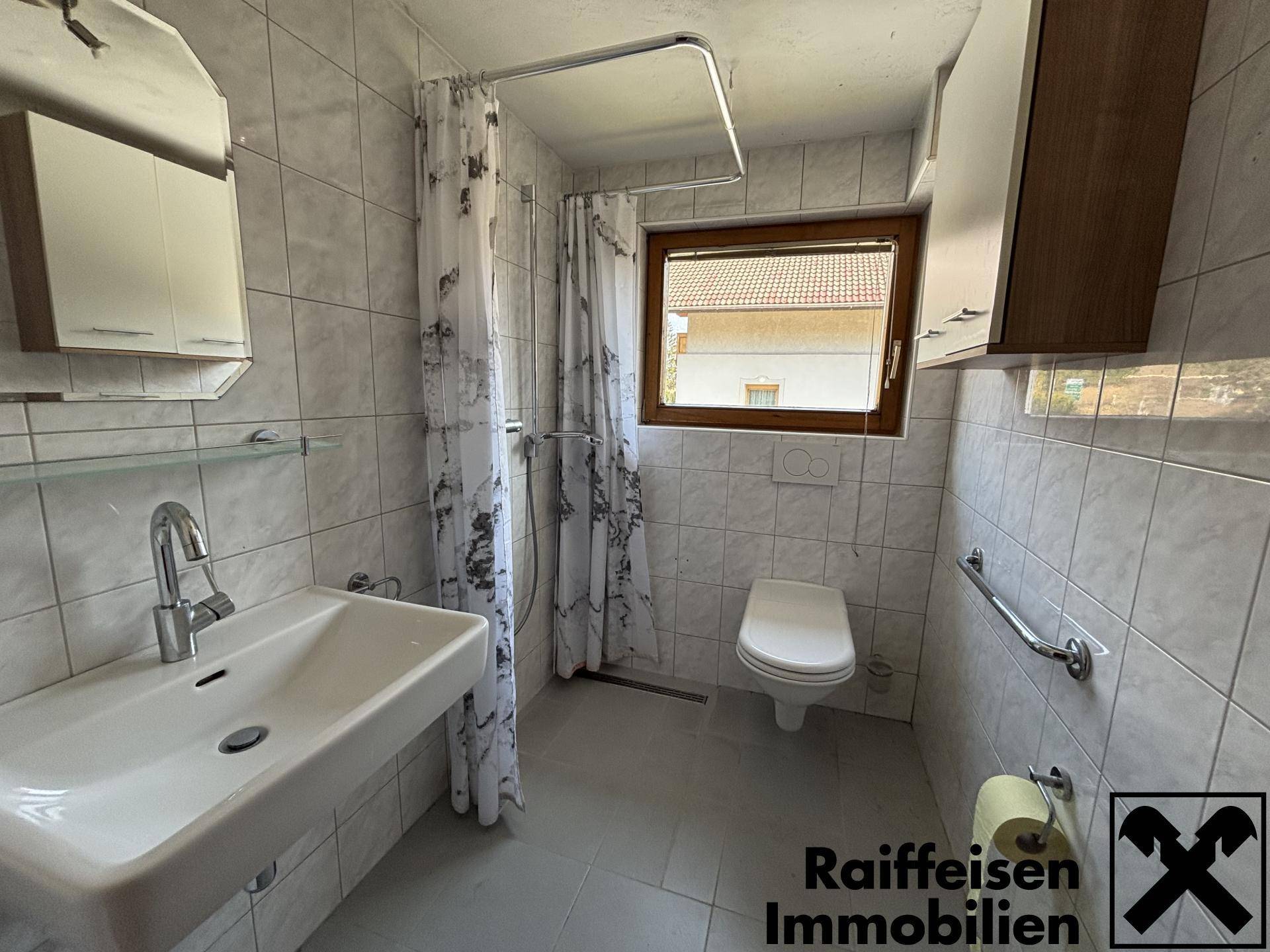 Gefliestes Badezimmer mit Dusche, WC, Einzelwaschbecken und Haltegriffen.