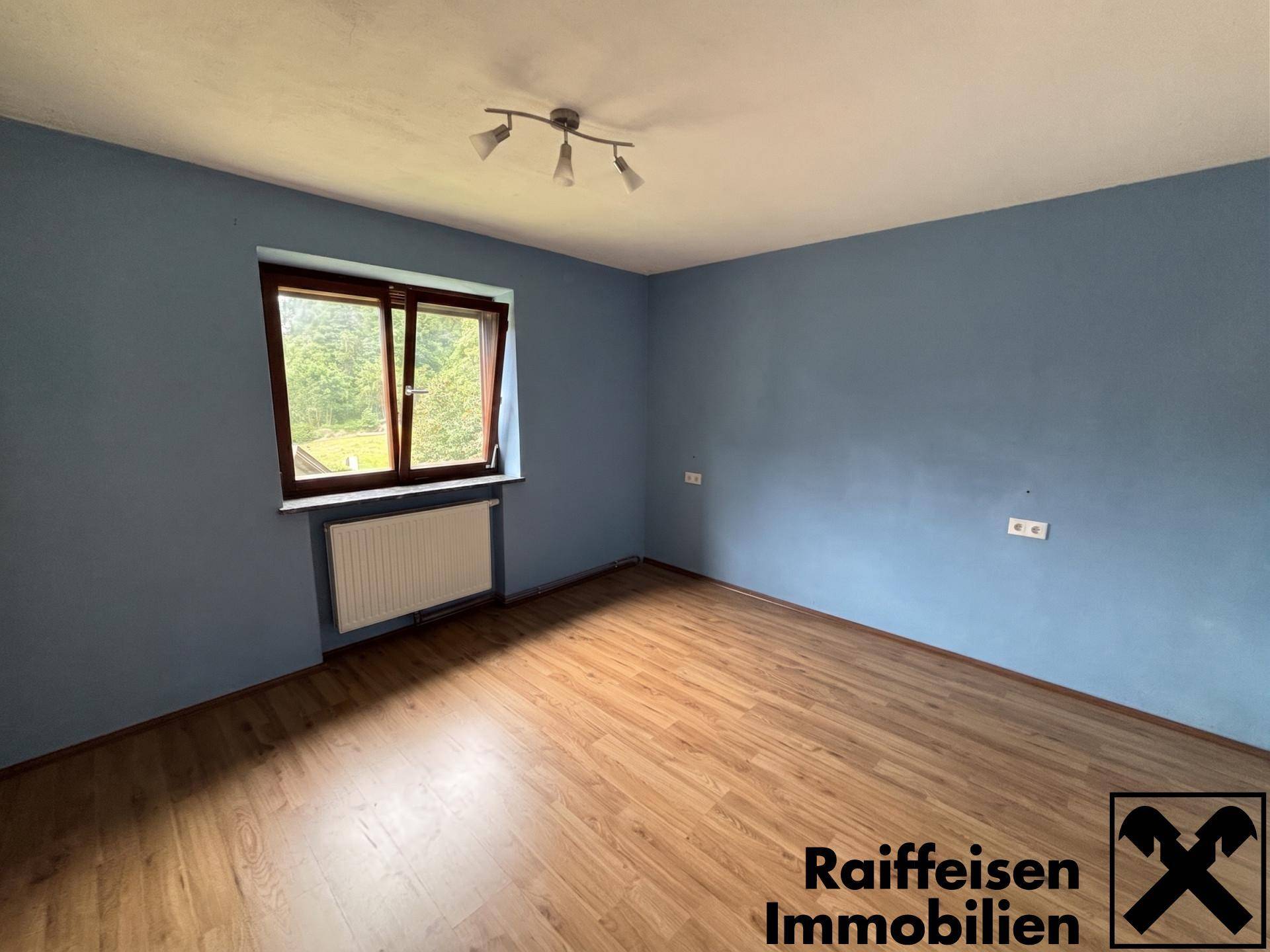 Zimmer mit blauer Akzentwand, Laminatboden und Fenster mit Blick auf die grüne Landschaft.