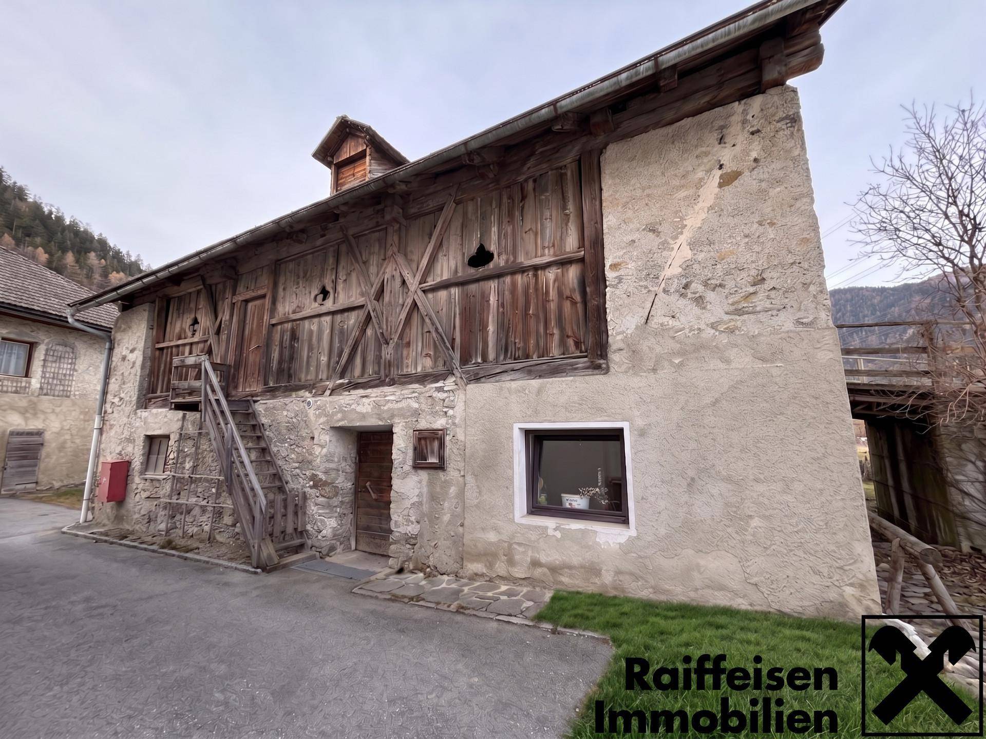 Altes Holzgebäude mit Außentreppe und Steinfassade, umgeben von ländlicher Umgebung.