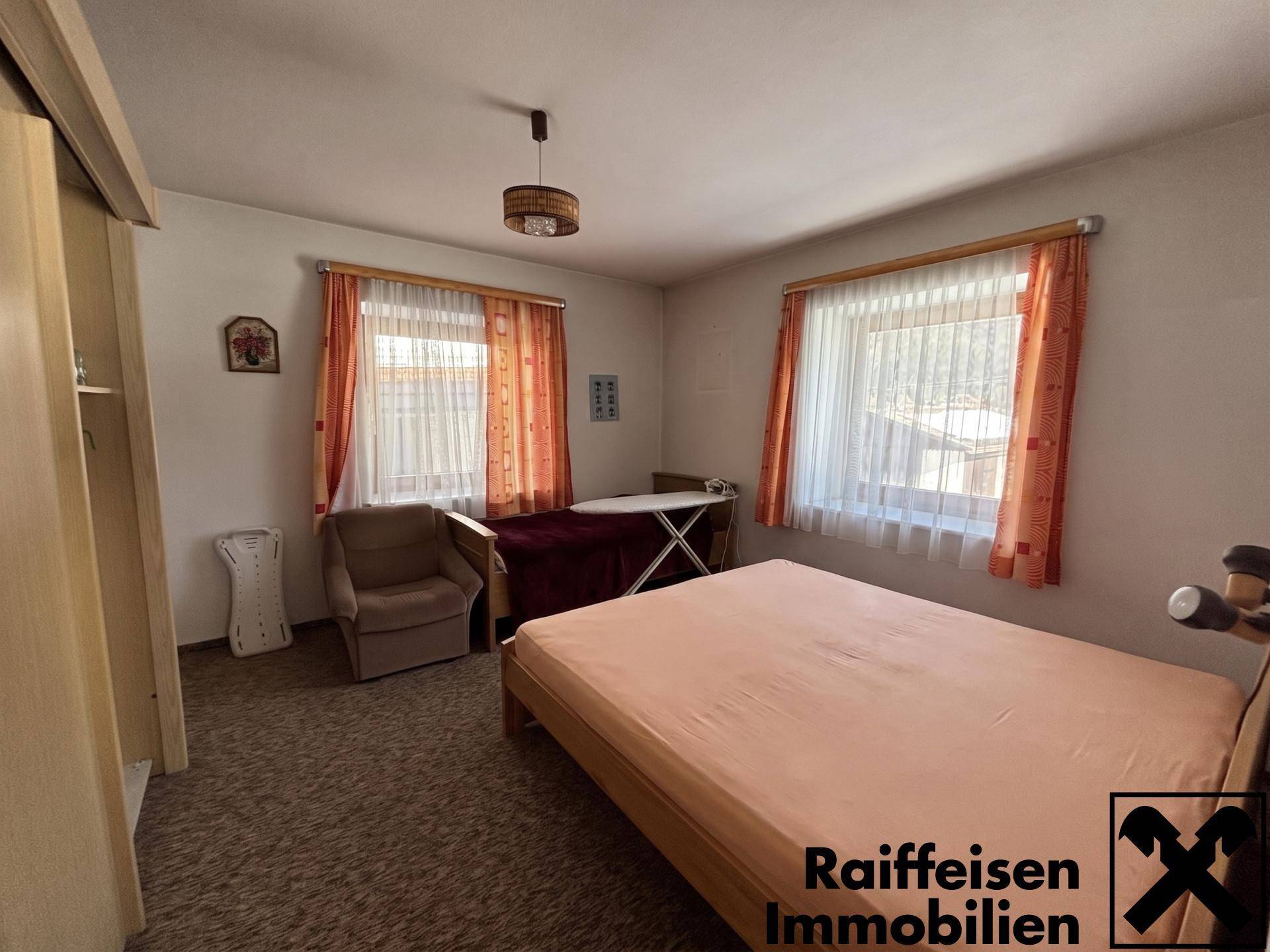 Schlafzimmer mit Doppelbett, Sessel und zwei Fenstern, die viel Tageslicht hereinlassen.