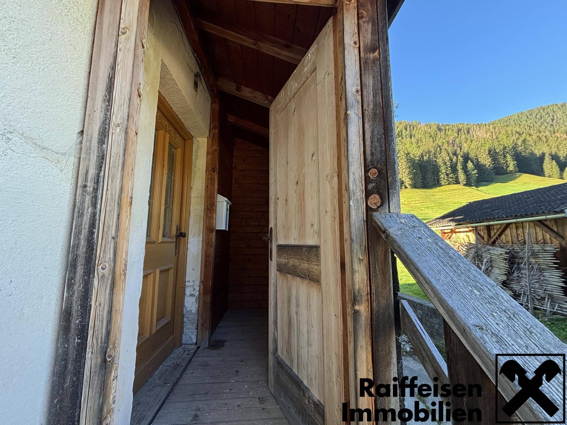 Rustikaler Hauseingang mit Holztür und Geländer, umgeben von grüner Natur.