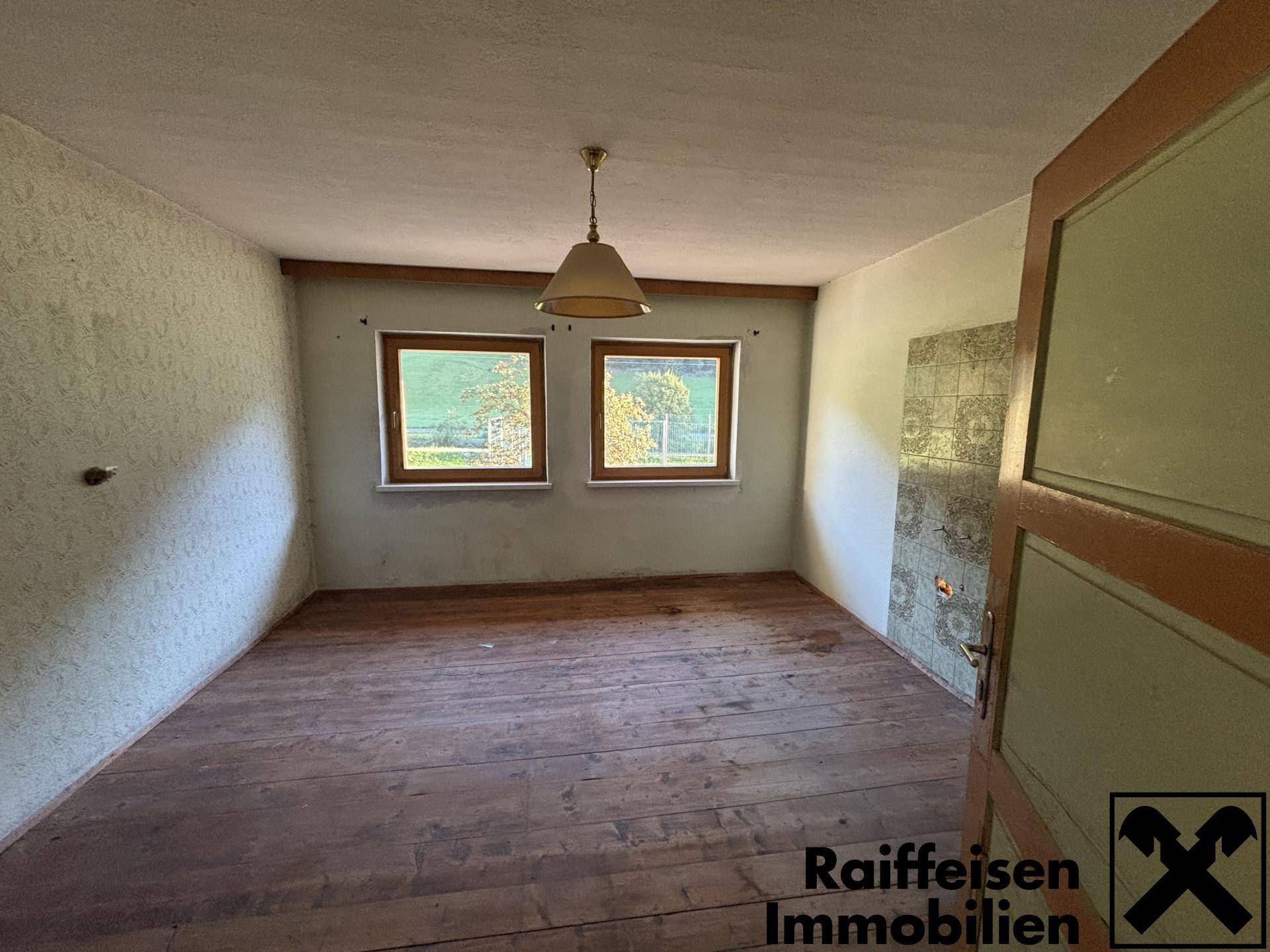 Helles Zimmer mit Holzdielenboden, zwei Fenstern und einem traditionellen Kachelofen.