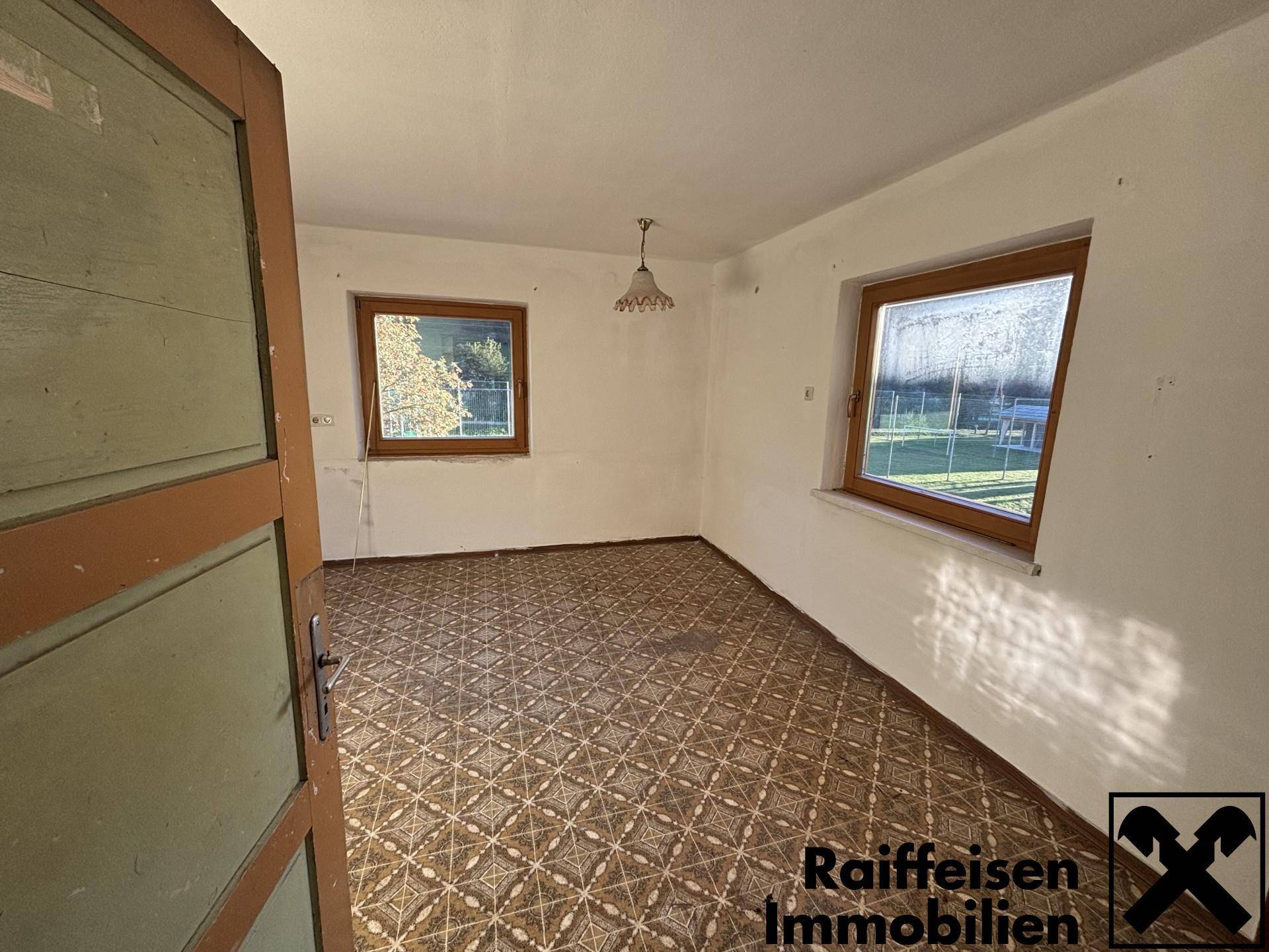 Leeres Zimmer mit gemustertem Fliesenboden, zwei Fenstern und Zugang zu weiteren Bereichen.