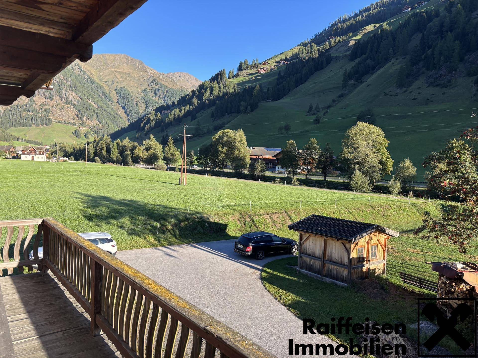 Aussicht vom Holzbalkon auf den Parkplatz mit kleinem Holzschuppen und die Bergkulisse.