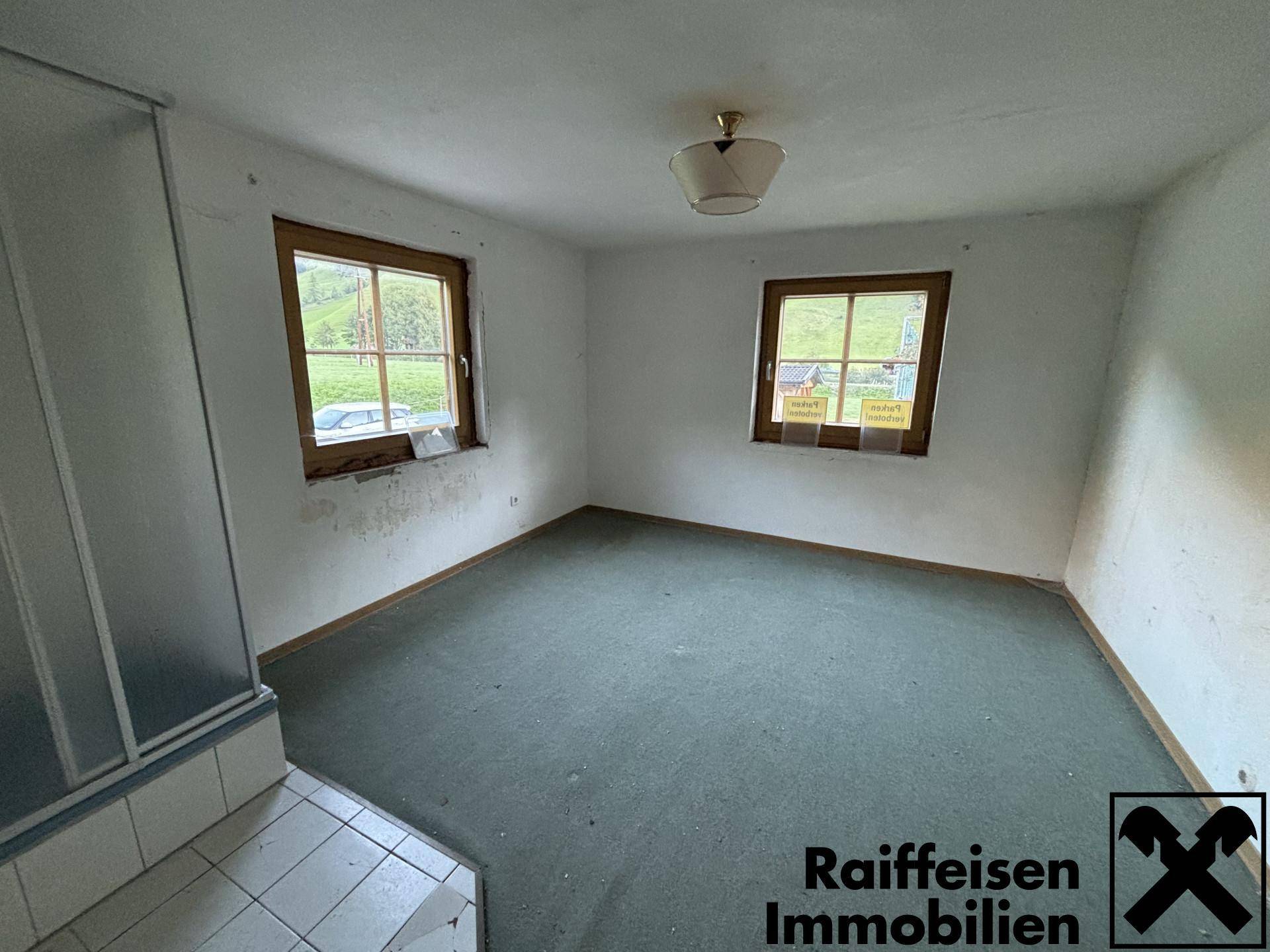 Leeres Zimmer mit grünem Teppichboden, zwei Fenstern und einer integrierten Duschkabine.