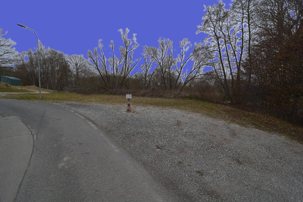 Unbefestigter Bereich mit Schotter und Gras, der zu einer Straße führt, umgeben von Bäumen.