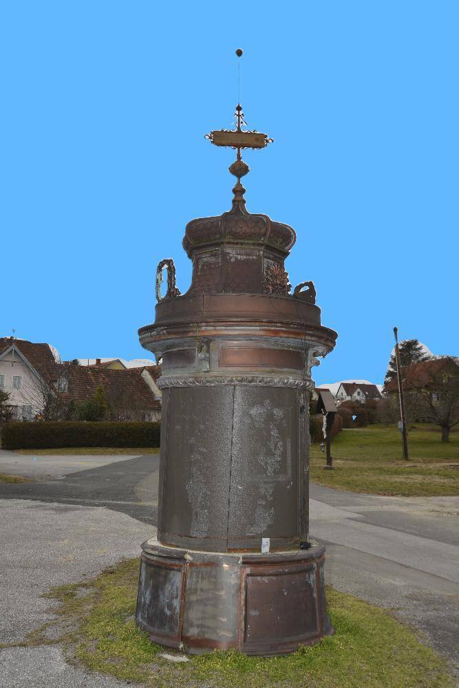 Nahaufnahme eines dekorativen Pfeilers mit Häusern und Grünflächen im Hintergrund unter blauem Himmel.