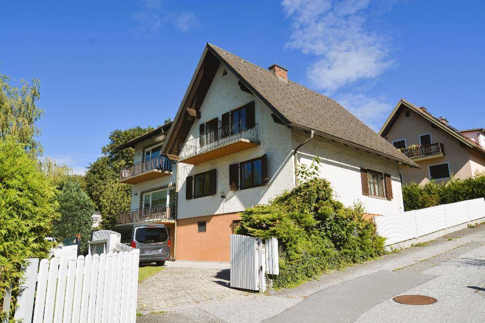 Ansicht des Hauses von der Straße mit weißem Zaun, Balkonen und umgebender Natur.