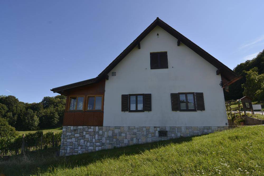 Seitenansicht des Hauses mit weißer Fassade, Steinsockel und Holzfensterläden, umgeben von Grünflächen.