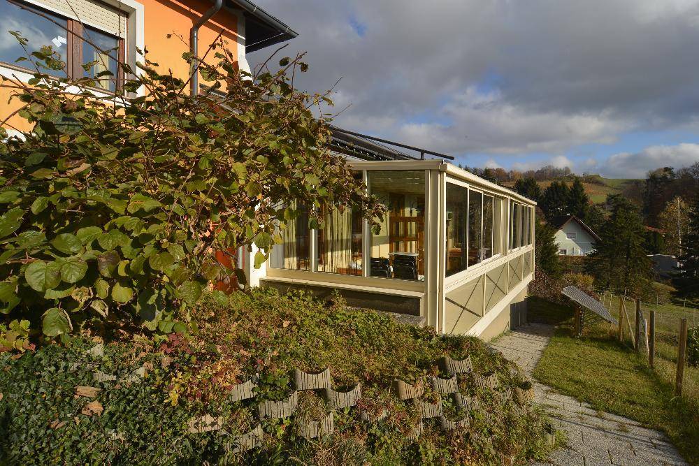 Seitenansicht des Hauses mit einem verglasten Wintergarten und üppiger Vegetation im Vordergrund.