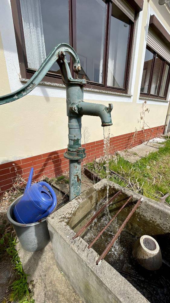 Alte Handschwengelpumpe, aus der Wasser in ein Betonbecken vor dem Gebäude fließt.