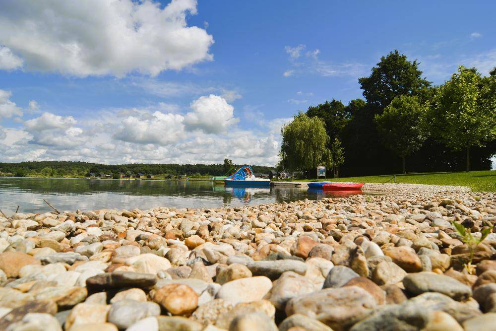 Kiesstrand am Ufer eines ruhigen Sees unter blauem Himmel mit weißen Wolken und grünen Bäumen.