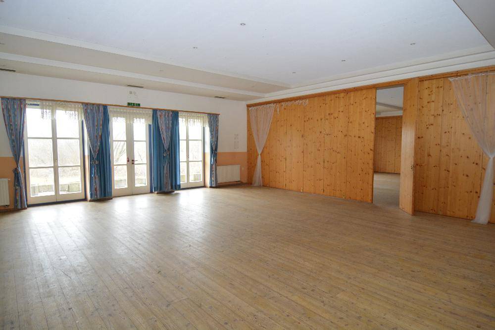 Großer leerer Saal mit Holzboden, Holzvertäfelung und Fenstertüren.