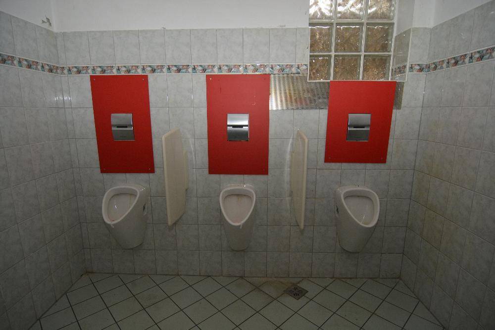 Öffentlicher Toilettenbereich mit drei Urinalen, roten Trennwänden und hellen Fliesen.