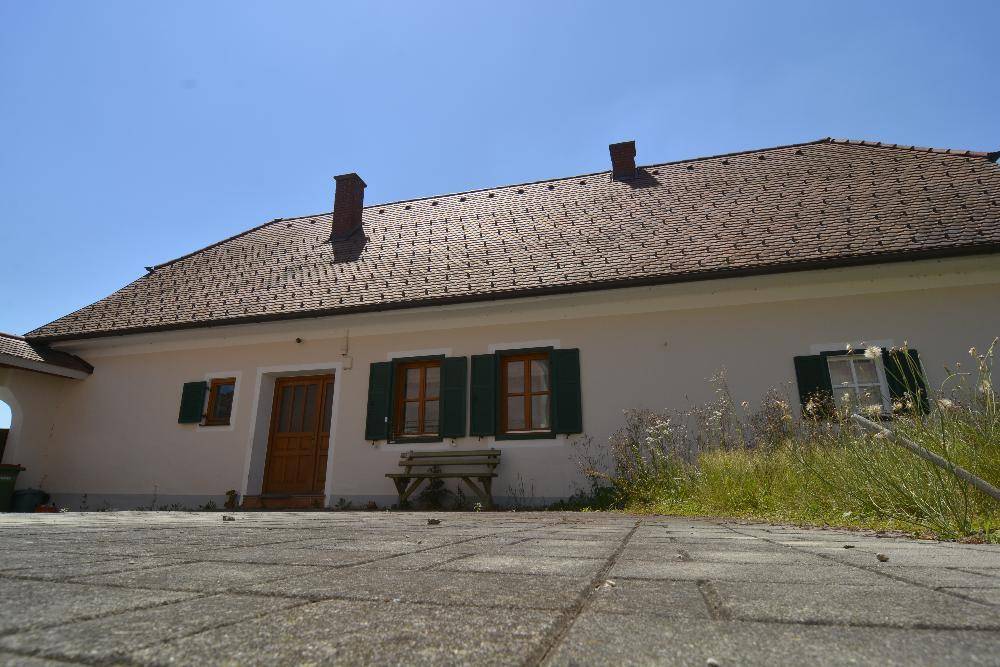 Nahaufnahme des Hauseingangs mit Holztür, grünen Fensterläden und gepflastertem Vorplatz.