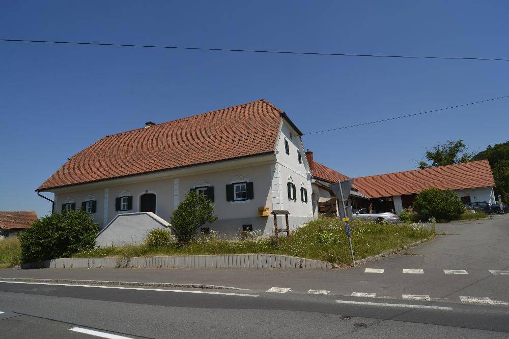 Traditionelles Landhaus mit Ziegeldach und grünen Fensterläden an einer ländlichen Straße.