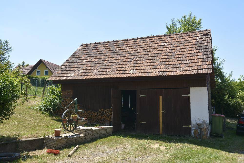 Hölzernes Nebengebäude mit Doppeltüren und Ziegeldach, umgeben von Grünflächen.