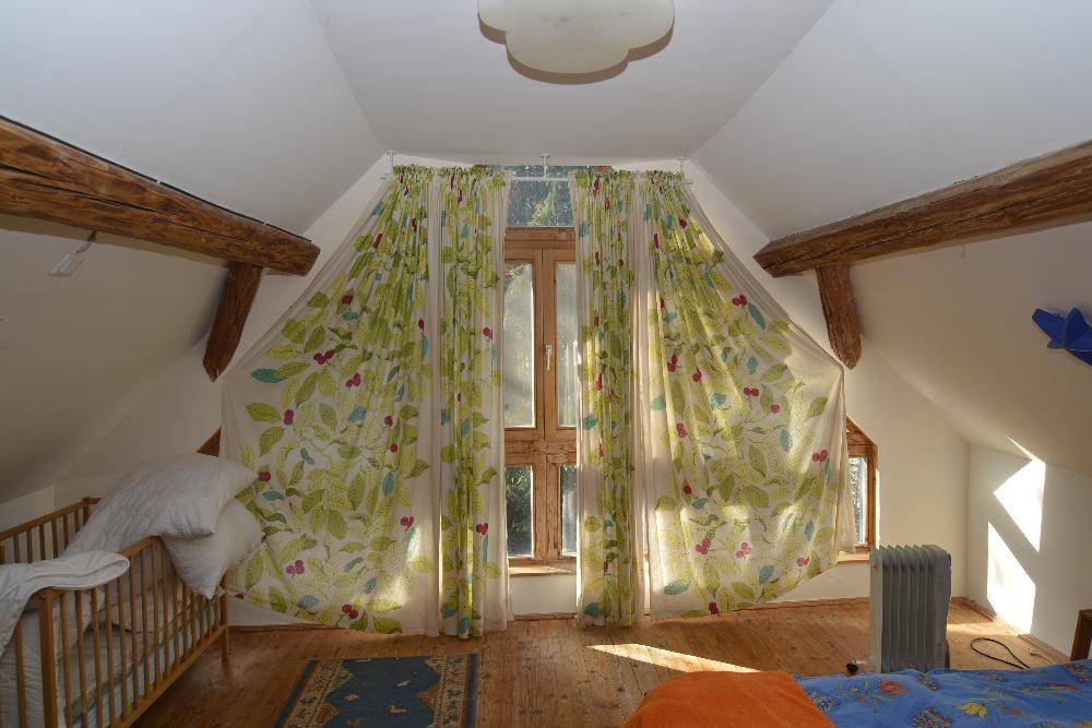 Helles Schlafzimmer im Dachgeschoss mit großen Fenstern, Holzbalken und Kinderbett.