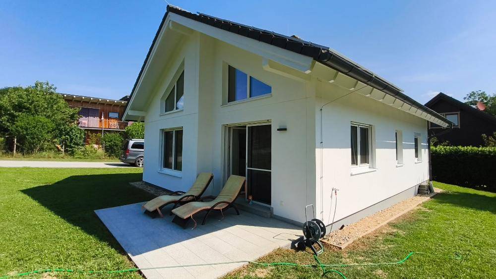 Modernes Einfamilienhaus mit weißer Fassade, großer Terrasse und Liegestühlen im sonnigen Garten.