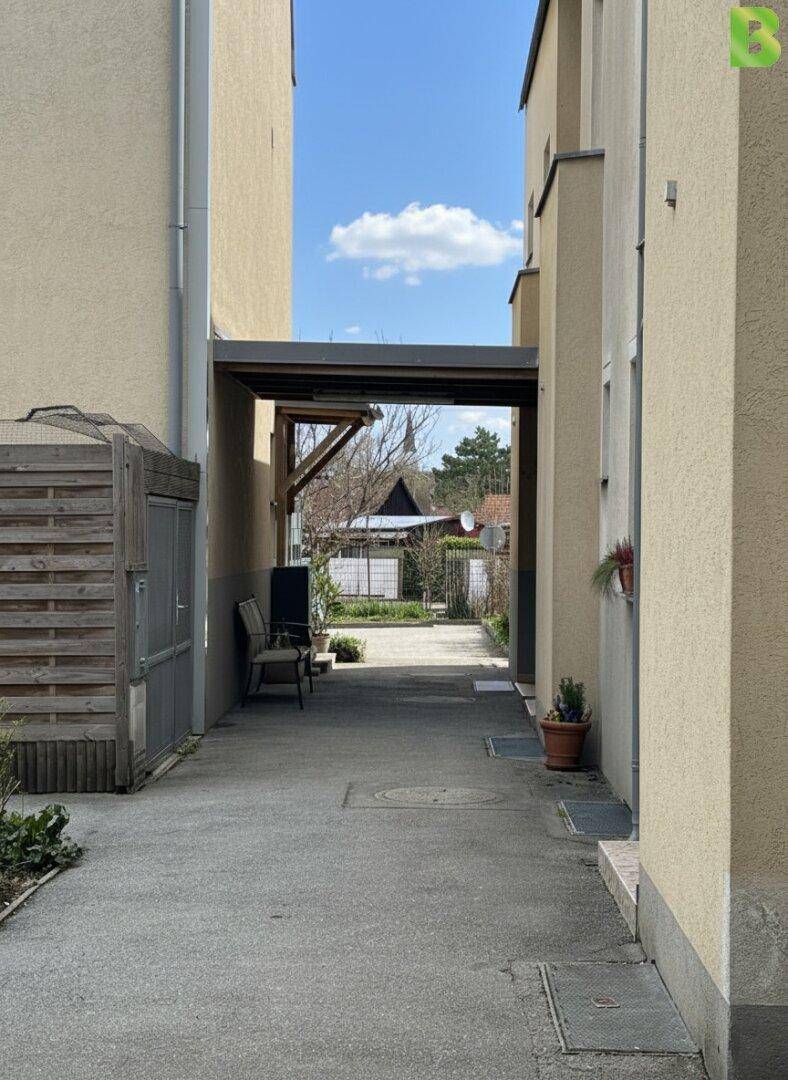 Durchgang zwischen Gebäuden mit Carport-Struktur und Blick auf die Umgebung.