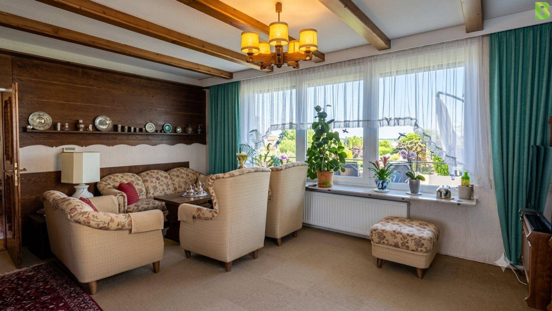 Gemütliches Wohnzimmer mit großen Fenstern, floralen Sesseln und Holzbalkendecke.