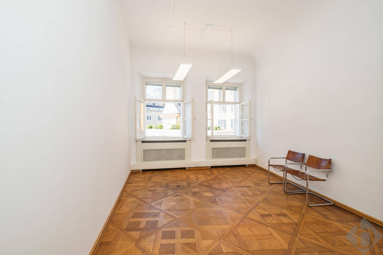 Heller Büroraum mit einzigartigem Parkettbodenmuster, zwei Fenstern und modernen Stühlen.