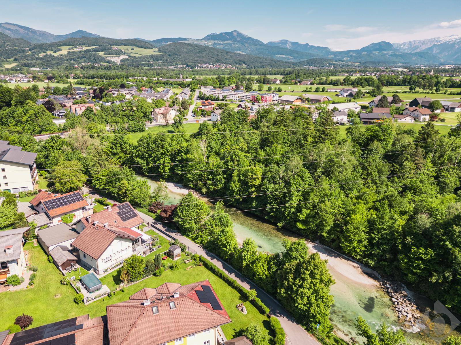 Weitläufige Luftaufnahme einer Siedlung mit Fluss, umgeben von grünen Hügeln und Bergen.