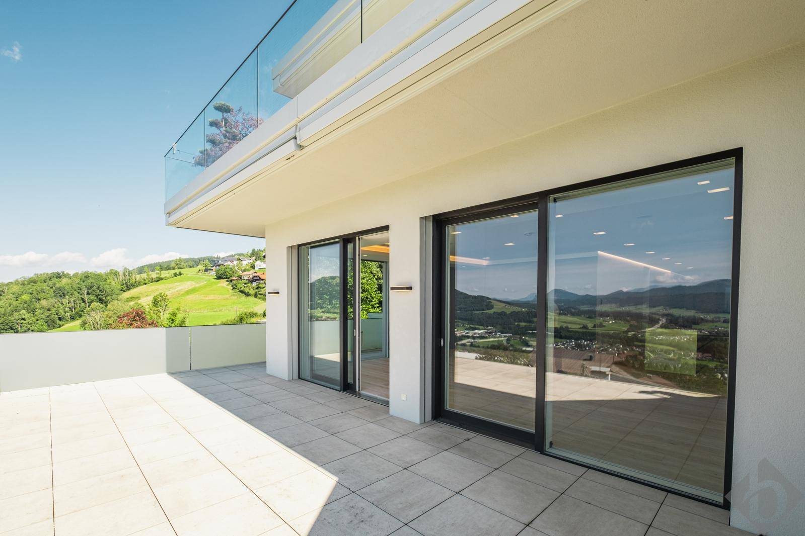 Geräumige Terrasse mit Fliesenboden, Glasgeländer und weitem Blick über grüne Hügel.