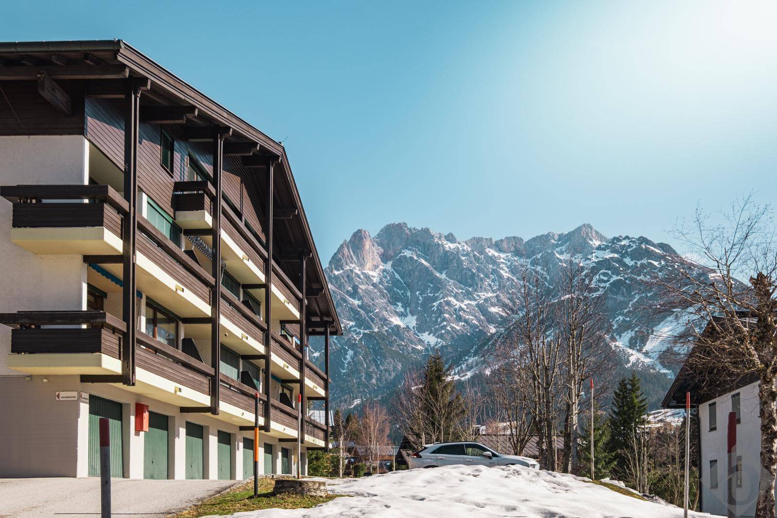 Mehrstöckiges Gebäude mit Balkonen und Garagen, umgeben von einer verschneiten Berglandschaft.