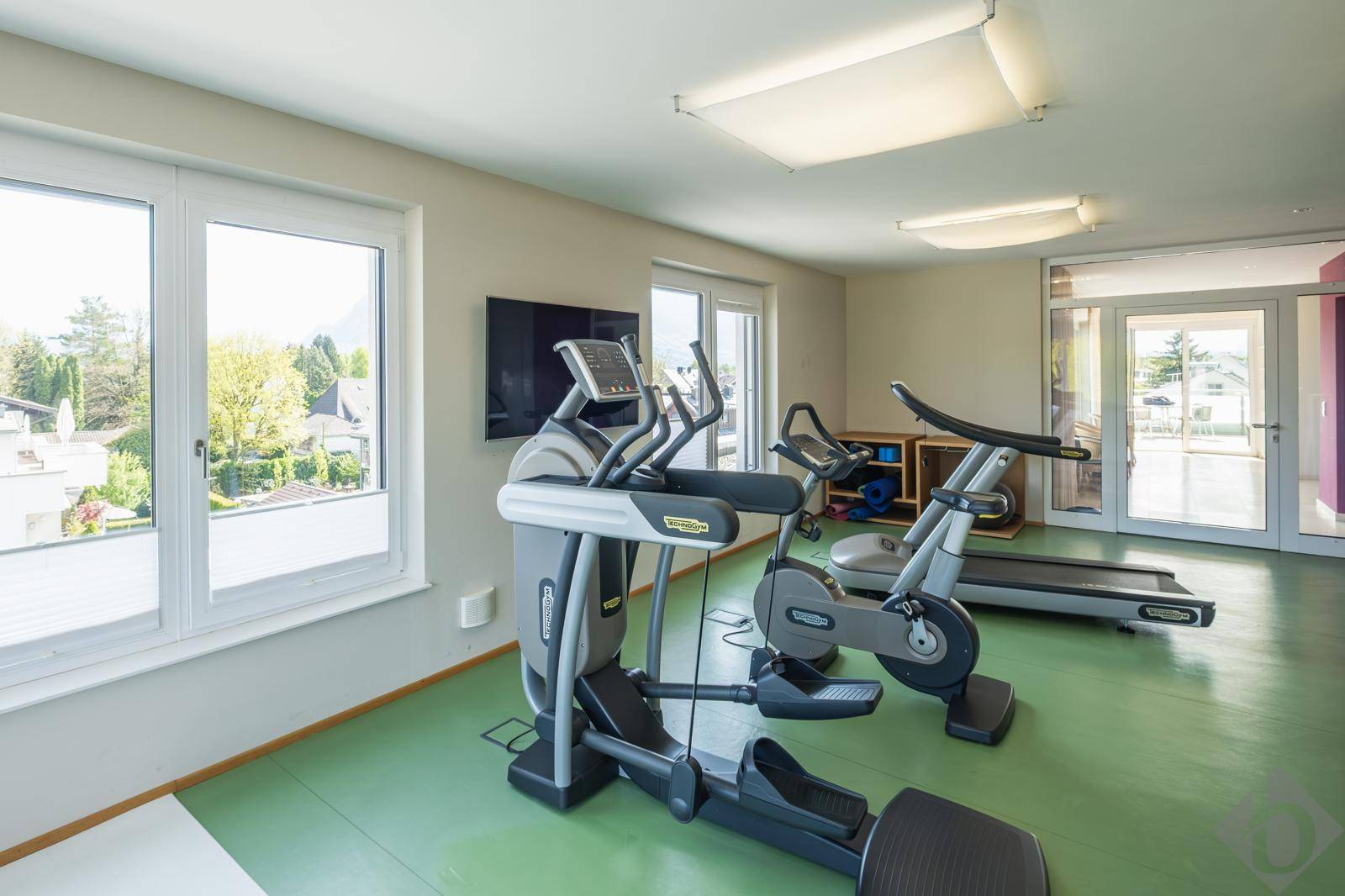 Gut ausgestatteter Fitnessraum mit verschiedenen Geräten und viel Tageslicht.