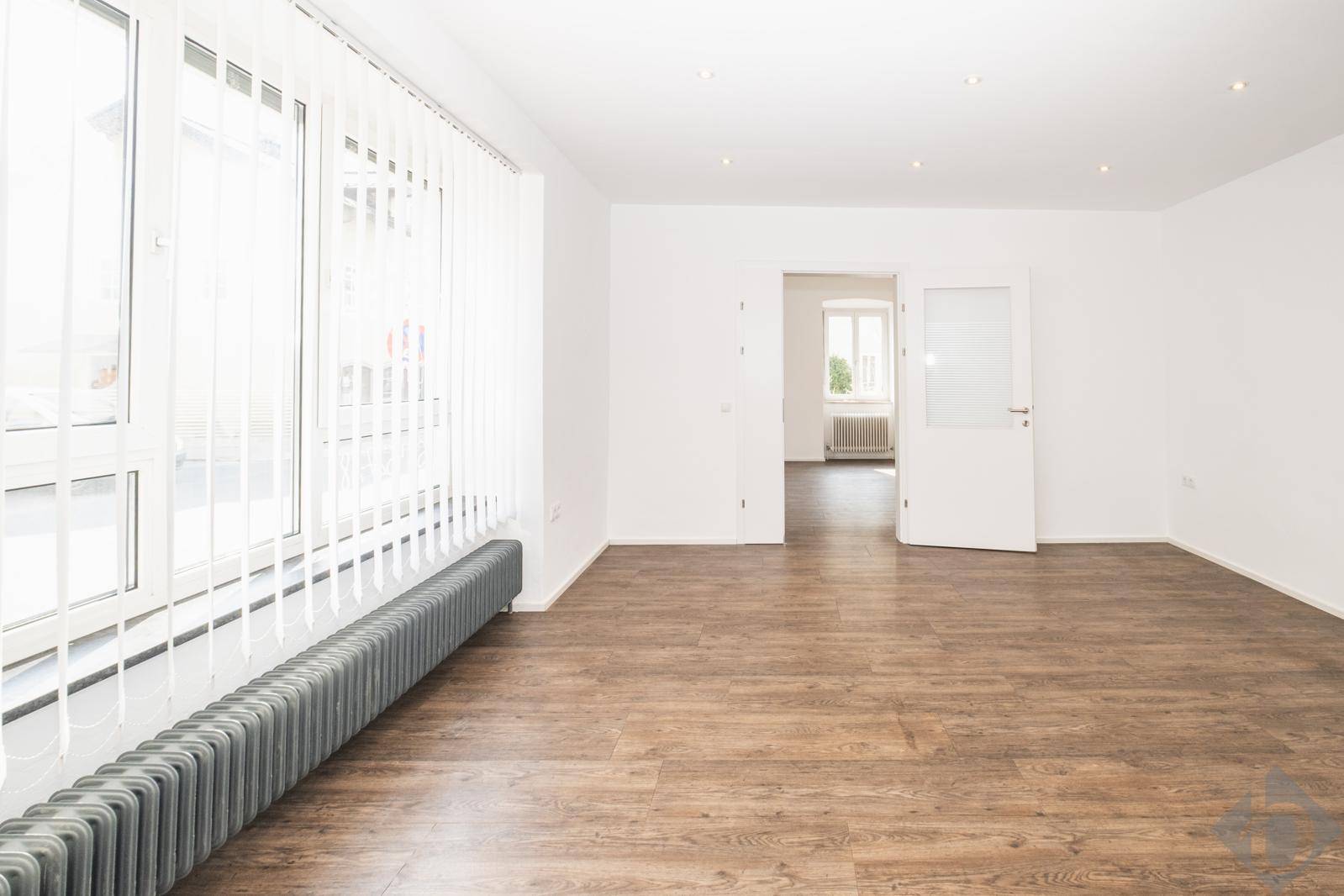 Heller Raum mit großen Fenstern, Laminatboden und Zugang zu einem weiteren Zimmer.