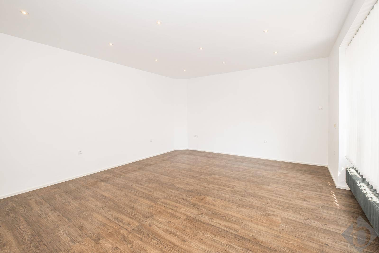 Leerer, heller Raum mit Laminatboden, Einbaustrahlern und einem Fenster mit Jalousien.