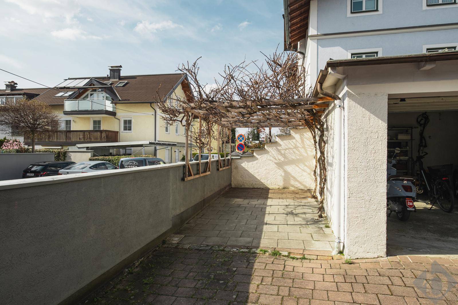 Gepflasterte Terrasse mit Pergola und angrenzender Garage, umgeben von Wohngebäuden.