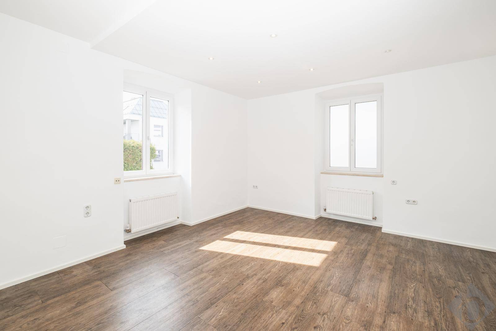 Heller Raum mit zwei Fenstern, Laminatboden und weiß gestrichenen Wänden.