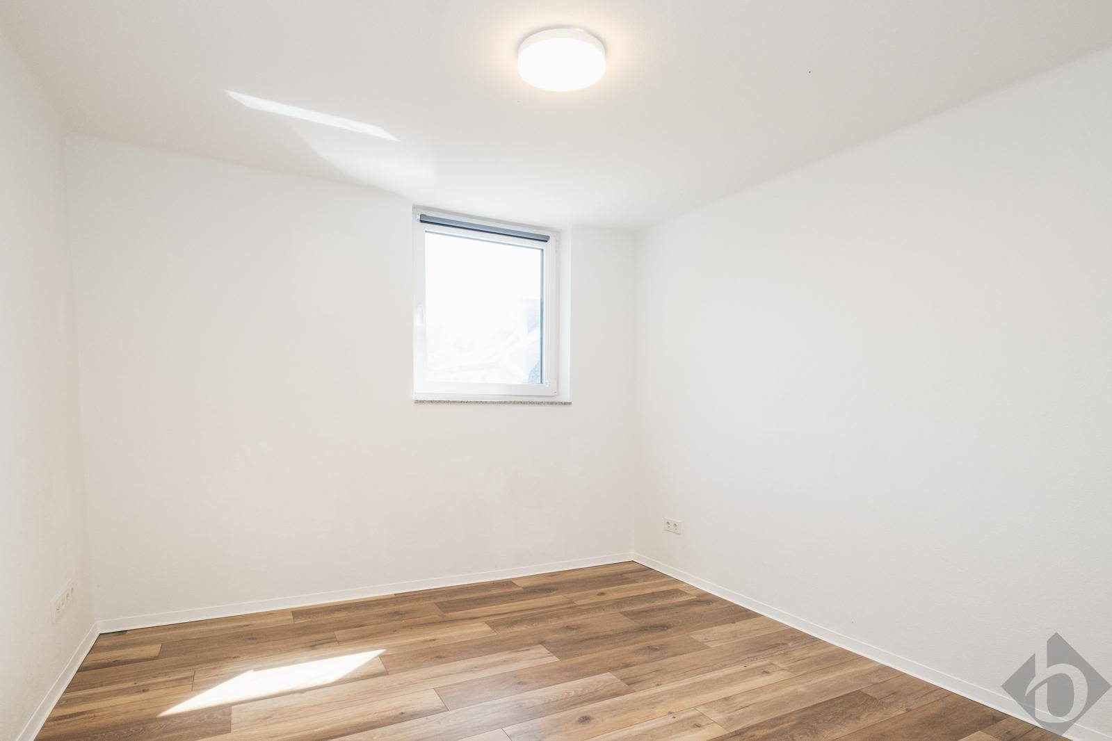 Leeres Schlafzimmer mit hellem Holzboden und einem Fenster, das viel Tageslicht hereinlässt.