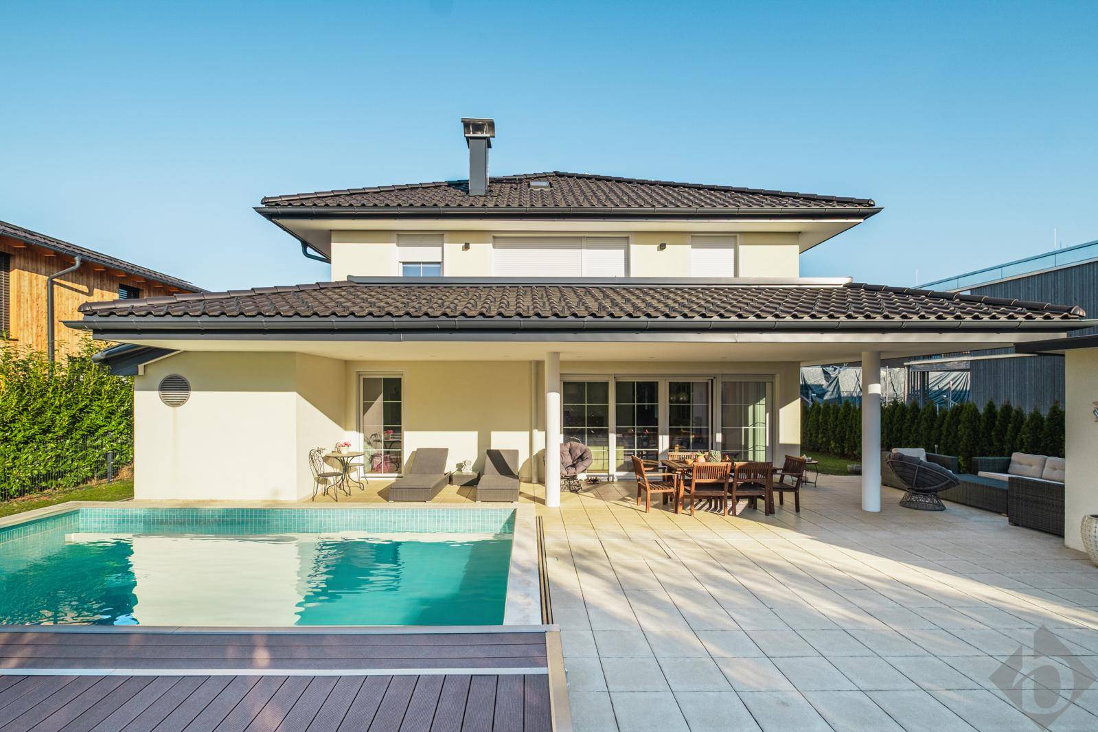 Rückansicht des Hauses mit großzügiger Terrasse, Swimmingpool und gepflegtem Gartenbereich unter blauem Himmel.