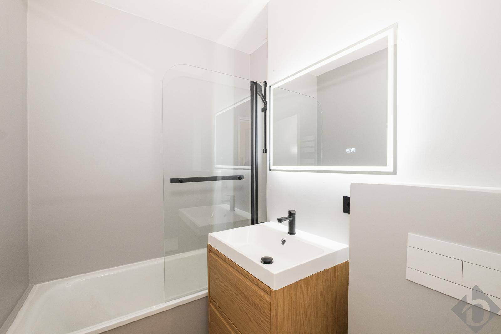 Modernes Badezimmer mit Waschtisch, beleuchtetem Spiegel, Badewanne mit Duschwand und Wand-WC.