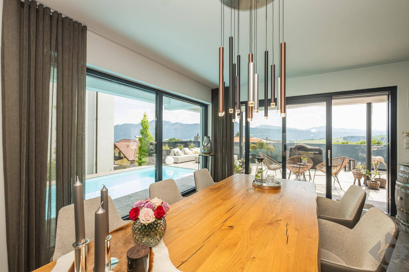 Heller Essbereich mit großem Holztisch, moderner Beleuchtung und direktem Zugang zur Poolterrasse mit Bergblick.