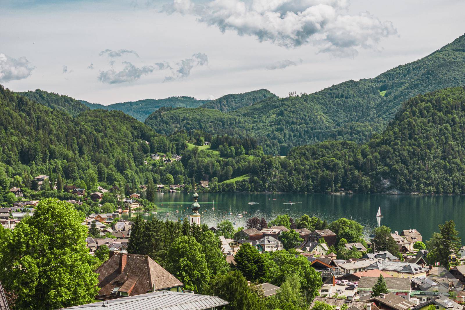 Weitläufiger Blick auf ein charmantes Dorf am See mit Kirchturm, umgeben von bewaldeten Hügeln.