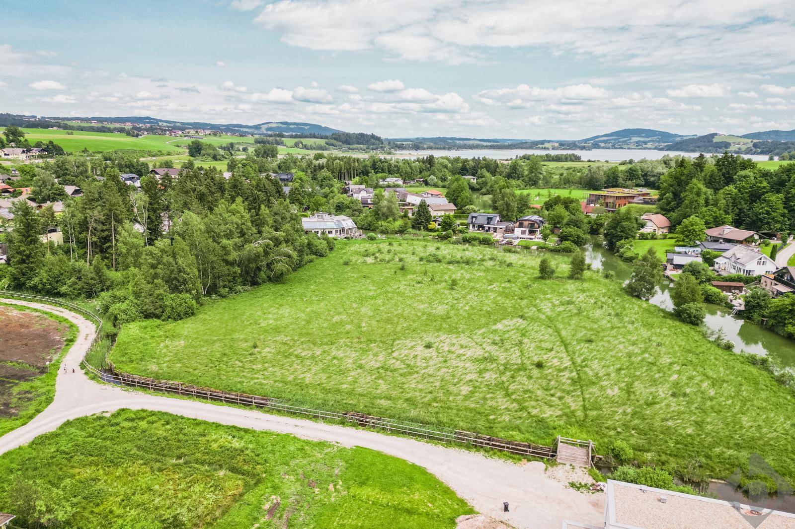 Weitläufige grüne Landschaft mit einem See im Hintergrund und einer kleinen Brücke im Vordergrund.