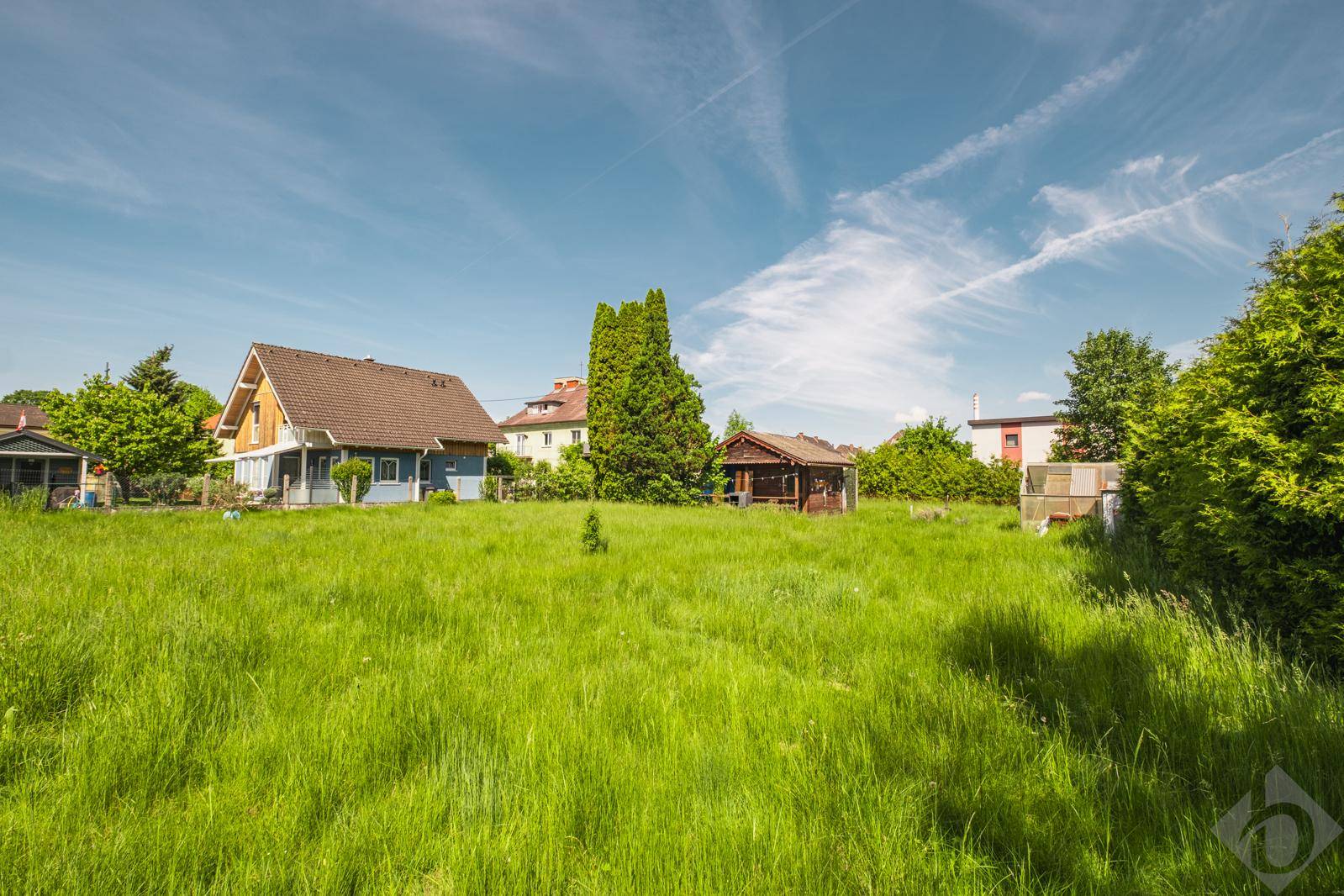 Weitläufiges grünes Grundstück mit einem kleinen Holzhaus und umliegenden Wohnhäusern unter blauem Himmel.