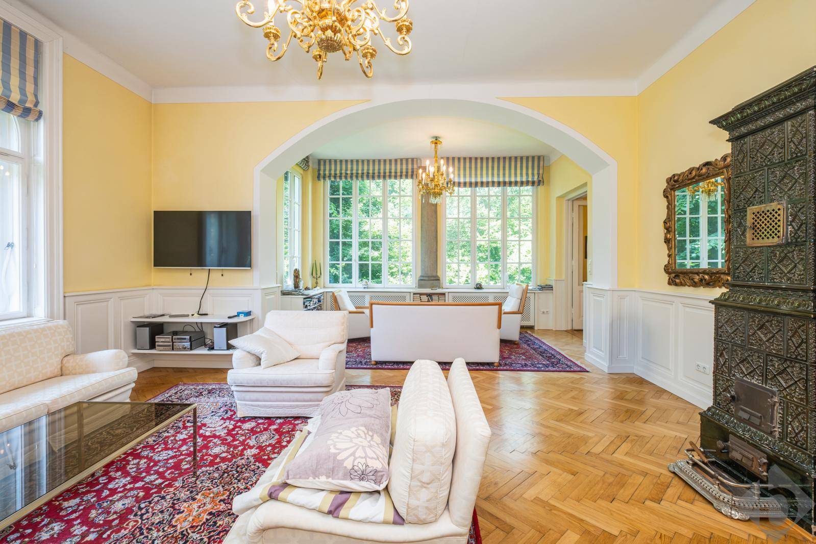 Großzügiges Wohnzimmer mit Parkettboden, Kachelofen und großen Fenstern mit Blick ins Grüne.