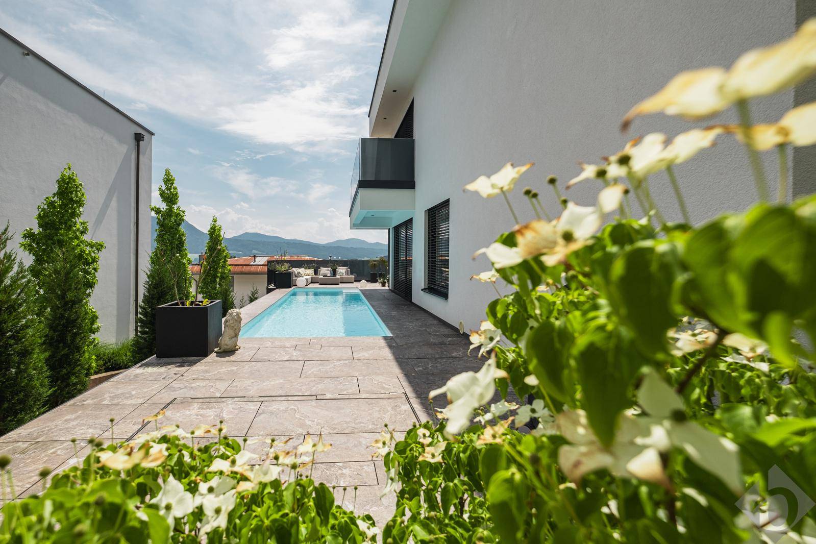 Seitlicher Blick auf die moderne Villa mit Poolbereich, Terrasse und blühenden Pflanzen im Vordergrund.