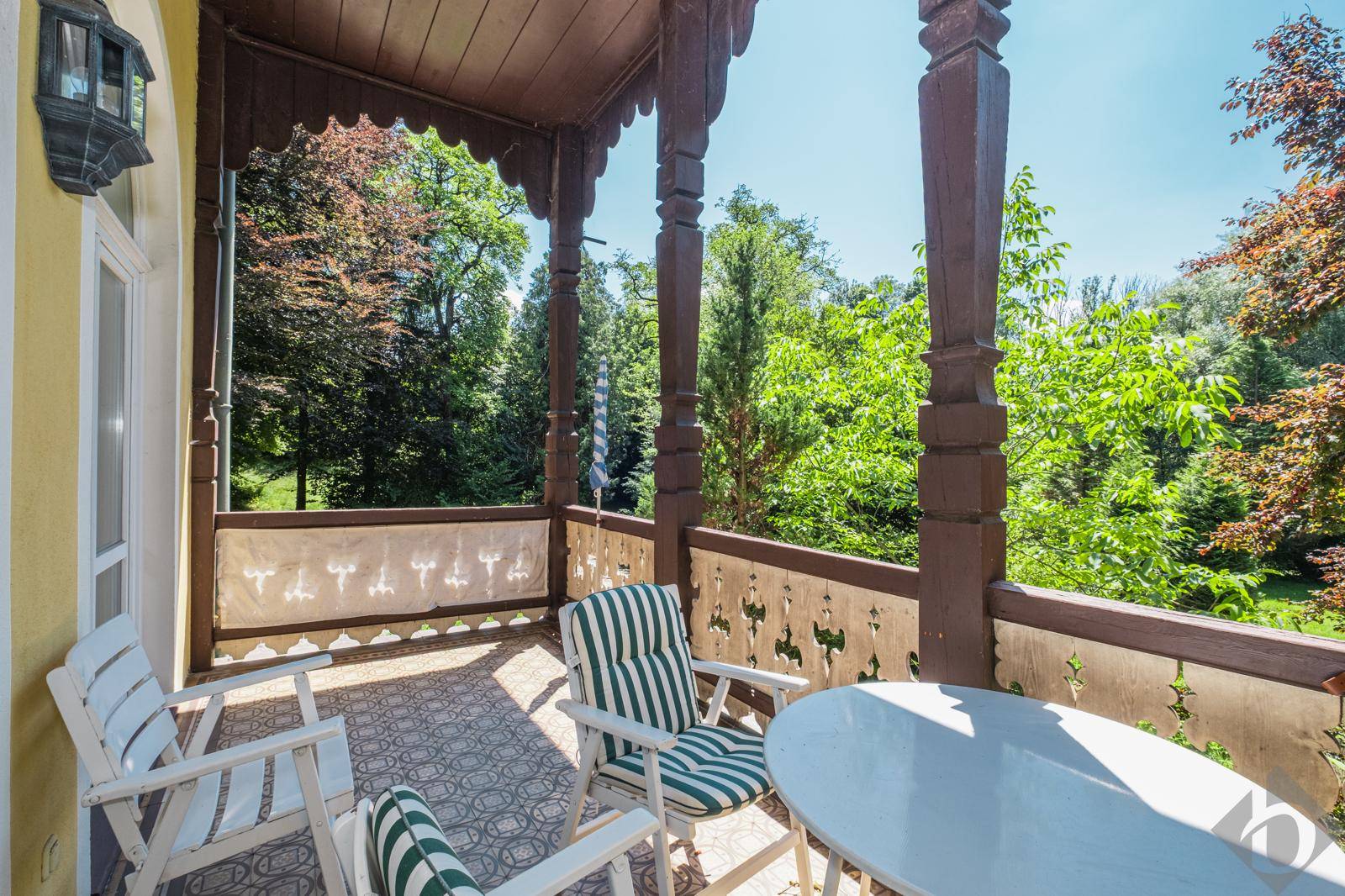 Ein sonniger Balkon mit Holzsäulen und Gartenmöbeln, umgeben von üppigem Grün und Bäumen.