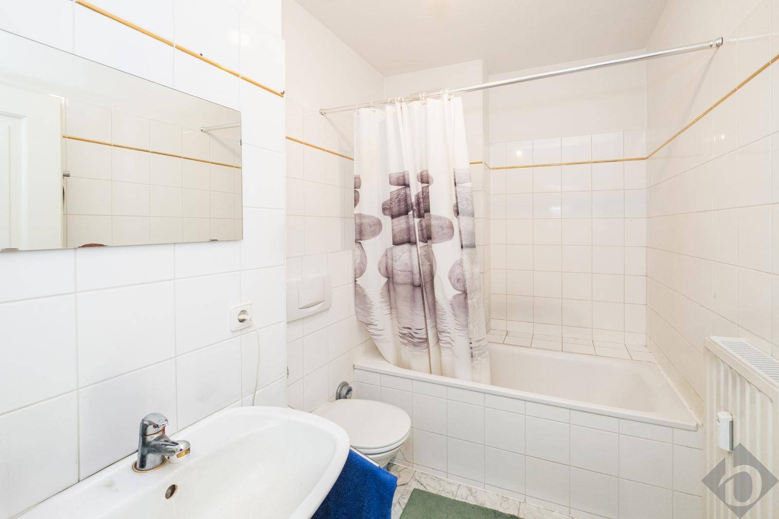 Helles Badezimmer mit weißen Fliesen, Badewanne mit Duschvorhang und einem einzelnen Waschbecken.