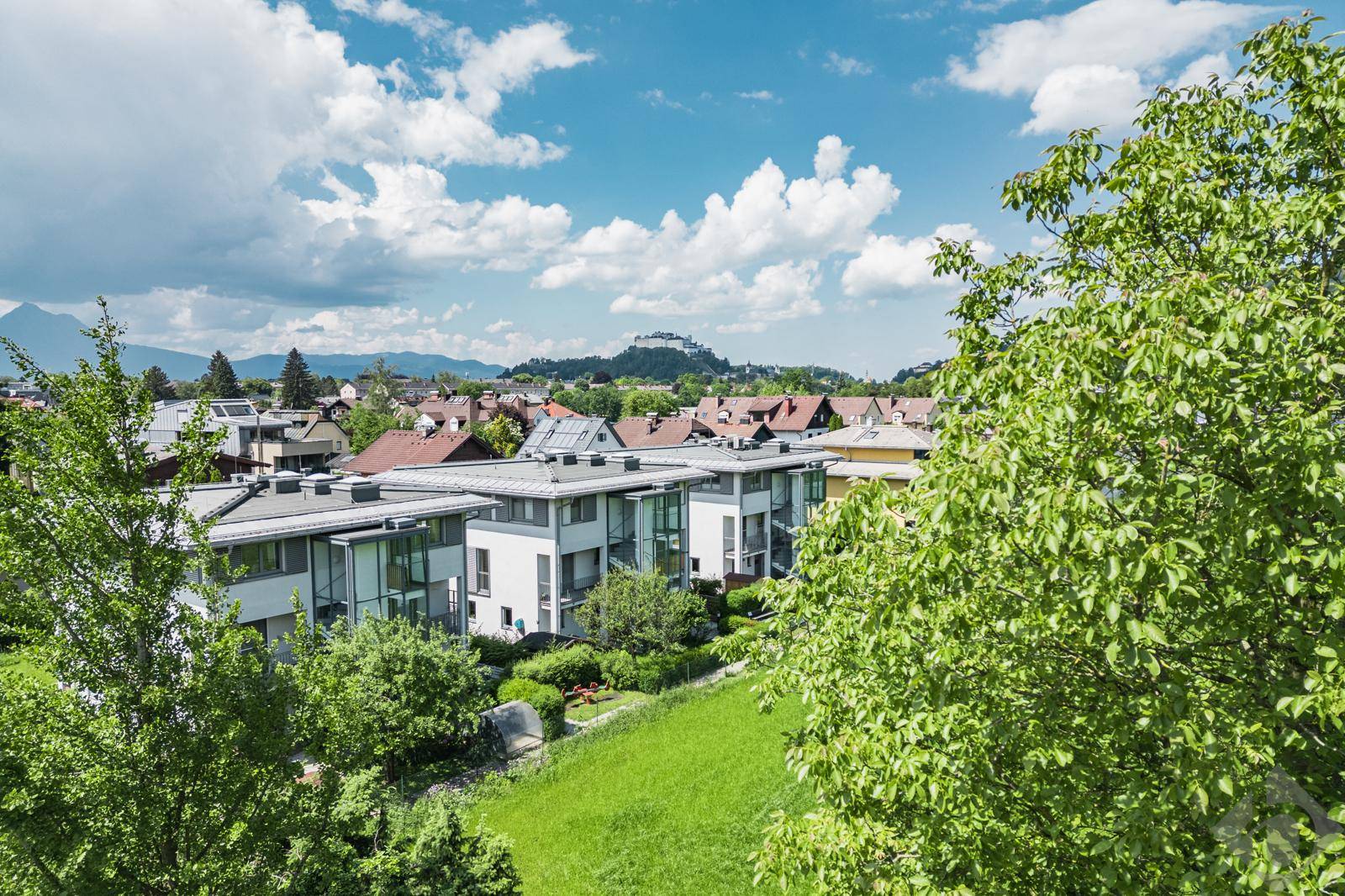 Panoramablick über eine Wohnsiedlung mit viel Grün und Bergen im Hintergrund unter blauem Himmel.